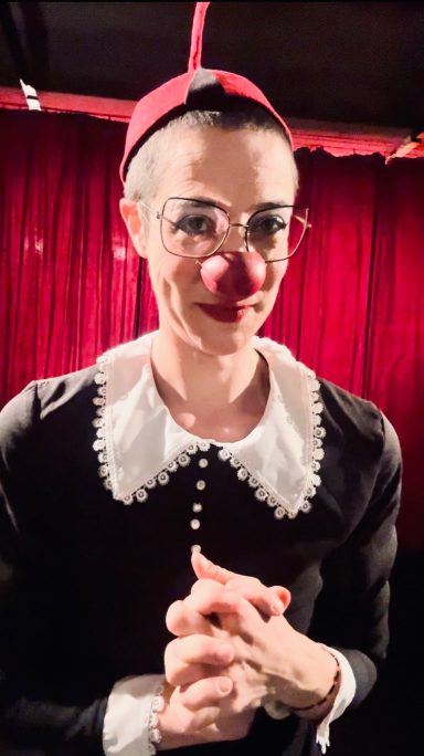 Titania Chaos looking witty in a black and white dress on a stage with a red curtain in Theater Ole in Vienna. 